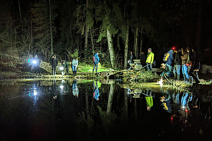 Eine Menschengruppe läuft im Dunkeln an einem Gewässer entlang.