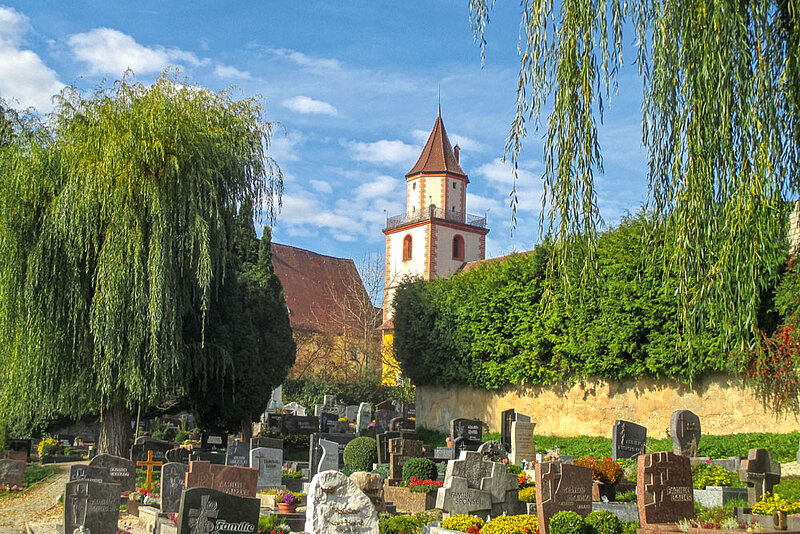 Grüner Friedhof mit Kirchturmspitze im Hintergrund.