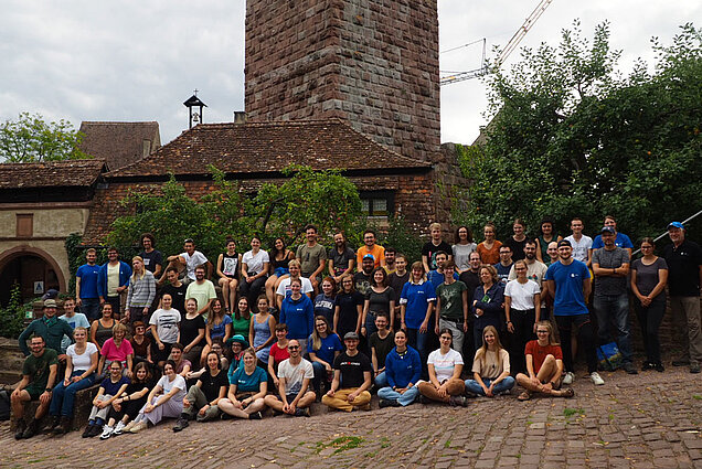 Eine große Gruppe von Jugendlichen sitzt und steht vor einem mittelalterlichen Turm.
